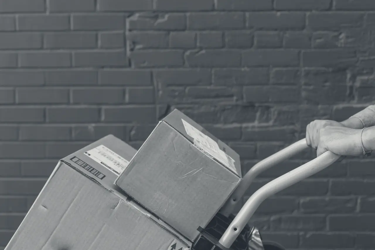Cardboard boxes on a hand truck being pushed past a brick wall.